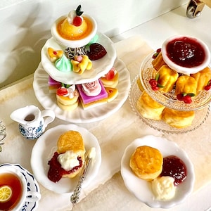 Puede incluir: Un juego de té de la tarde en miniatura con una bandeja de servicio de varios niveles con postres, que incluyen pasteles, bizcochos y natillas. Los bollos con mermelada y crema están en platos pequeños, junto con una taza de té con una rodaja de limón. También hay una cremera en miniatura.
