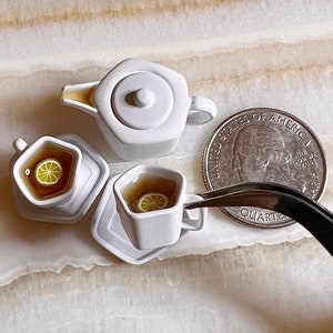 May include: A miniature white teapot, two teacups, and saucers with a lemon slice in each cup. A US quarter is included for scale.