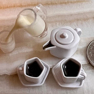 May include: A white teapot with a lid, two white cups with saucers, and a clear pitcher with a white liquid pouring out. The cups and saucers are shaped like a diamond. The teapot and cups are on a light brown surface.