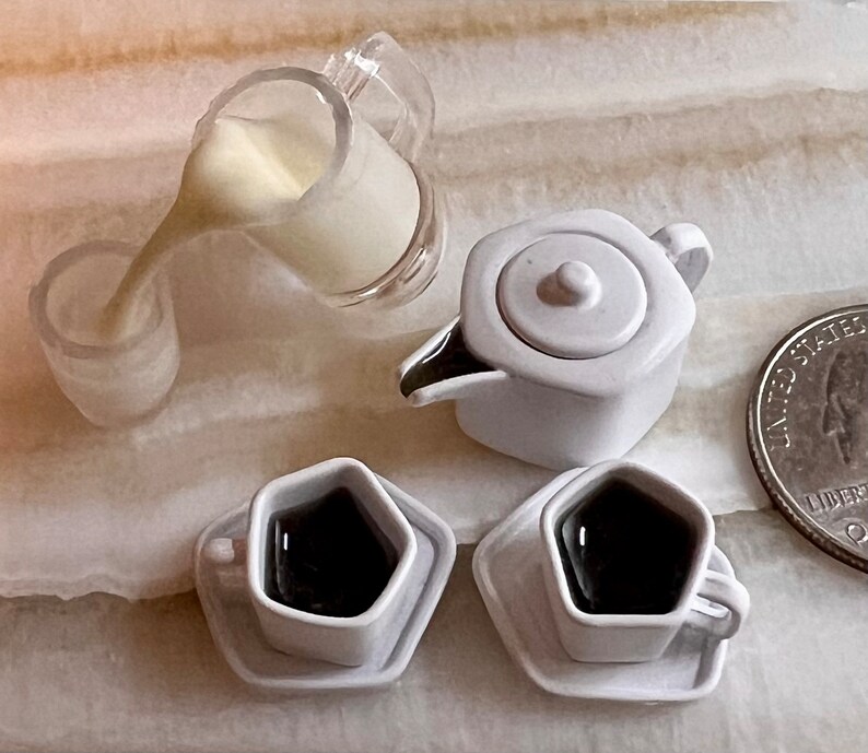 May include: A white teapot with a lid, two white cups with saucers, and a clear pitcher with a white liquid pouring out. The cups and saucers are shaped like a diamond. The teapot and cups are on a light brown surface.