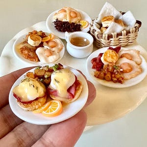 Op de afbeelding: Miniatuur ontbijtgerechten op witte borden, waaronder Eggs Benedict, gebakken eieren en gebak. Een klein mandje bevat biscuits. Er zijn ook sinaasappelschijfjes en een klein wit kopje saus. Het eten is gerangschikt op een wit oppervlak.