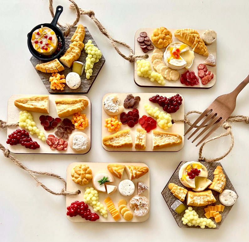 May include: Six miniature wooden cheese boards with various toppings, including cheese, crackers, grapes, and fruit. The boards are decorated with red and white accents and are arranged on a white surface.