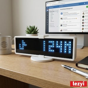 Puede incluir: Un reloj digital blanco que muestra la hora 12:44 en luces LED azules. El reloj está sobre un soporte blanco en un escritorio de madera. Una taza con la palabra "Followers" y un monitor de computadora están en el fondo.