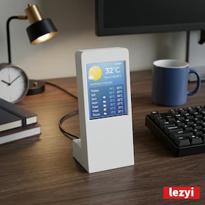 Puede incluir: Una estación meteorológica blanca muestra un pronóstico de siete días con temperaturas en grados Celsius. La pantalla muestra 32°C y sensación térmica de 36°C. También se ven una lámpara de escritorio, un reloj, un cuaderno, una taza y un teclado.