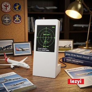 Op de afbeelding: Een wit digitaal radarscherm met een groene cirkelvormige radarweergave en tekst die de afstand en hoogte van vliegtuigen toont. Het apparaat staat op een houten bureau, omringd door boeken, modellen en foto's met een luchtvaartthema.