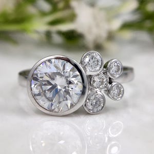 Può includere: Un anello d'argento con una grande gemma rotonda e trasparente in una montatura a castone, accompagnata da un gruppo di gemme rotonde più piccole. L'anello è ambientato su uno sfondo sfocato di fiori bianchi.