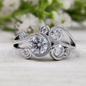 Könnte beinhalten: Ein silberner Ring mit einem großen runden Diamanten, der von kleineren Diamanten umgeben ist. Der Ring hat ein einzigartiges Design mit einem wellenförmigen Band.