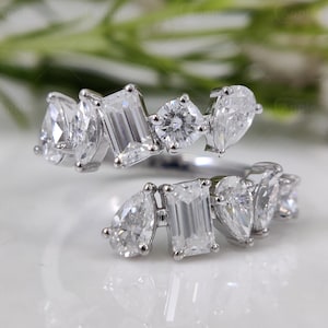 Può includere: Un anello d'argento con un gruppo di pietre preziose chiare e scintillanti di varie forme, tra cui a goccia, smeraldo e rotonde. Il design dell'anello è moderno ed elegante.
