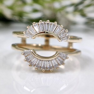 Op de afbeelding: Een goudkleurige ring met twee banden, elk met een halve cirkel van heldere baguette-geslepen stenen. Kleine, ronde accenten zijn langs de gebogen randen geplaatst. De ring is ontworpen om een andere ring te omvatten.
