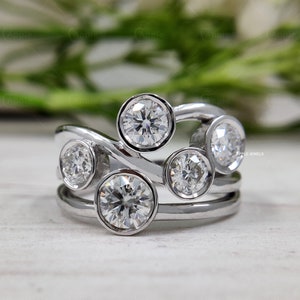 Könnte beinhalten: Ein silberner Ring mit fünf runden, klaren Edelsteinen, die in einem gebogenen Band gefasst sind. Der Ring liegt auf einer weißen Oberfläche.