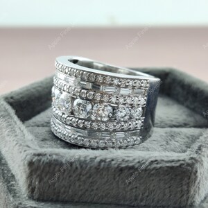 May include: A silver ring with multiple rows of round and baguette-shaped diamonds. The ring is set in a silver band and is displayed on a gray velvet box.