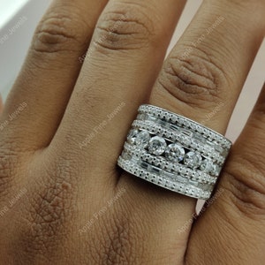 May include: A wide silver band ring with multiple rows of pave set diamonds. The ring is worn on a person's finger.