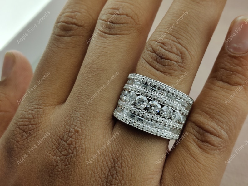 May include: A wide silver band ring with multiple rows of pave set diamonds. The ring is worn on a person's finger.