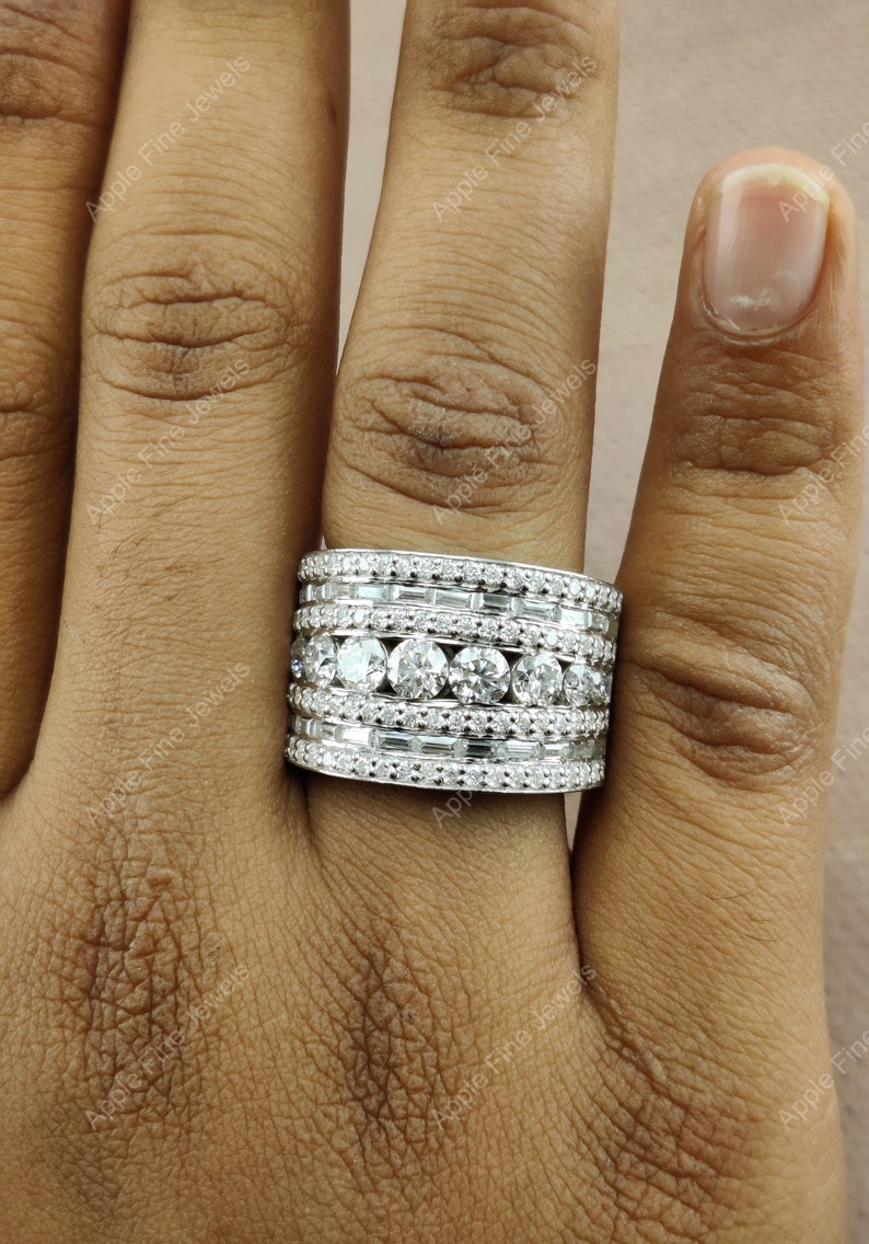 May include: A wide, silver band ring with multiple rows of sparkling diamonds. The ring is set with round and baguette-shaped diamonds.