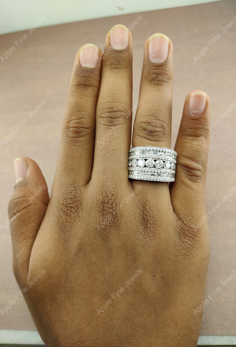 May include: A silver ring with multiple rows of round and baguette-shaped diamonds. The ring is on a person's finger.