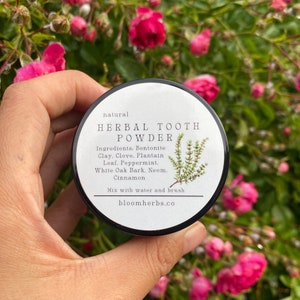 Può includere: Un barattolo bianco con un coperchio nero e testo nero che dice "natural HERBAL TOOTH POWDER Ingredients. Bentonite Clay. Clove. Plantain Leaf. Peppermint. White Oak Bark. Neem. Cinnamon Mix with water and brush bloomherbs.co"