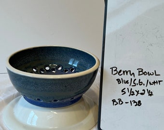 Op de afbeelding: Een blauwe en witte keramische bessenkom met een standaard. De kom heeft een blauwe glazuur met witte stippen en een witte rand. De standaard is wit. De kom heeft een diameter van 14 cm en een hoogte van 9 cm. De tekst op het witte bord luidt "Berry Bowl Blue/S.B./WHT 5 1/2 x 3 1/2 BB-138".
