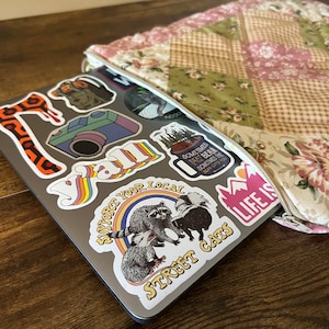 May include: A silver laptop with various colorful stickers on the lid. The stickers feature a rainbow, a raccoon, a skunk, a camera, a mug, a mountain, and the words "y'all", "support your local", "street cats", and "life is".