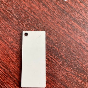 May include: A white rectangular tag with a small hole in the top left corner. The tag is made of a smooth, slightly textured material.