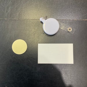 May include: A white retractable badge reel with a clear plastic strap and a metal clip. A round, yellow sticker and a white rectangular sticker are also present. The items are on a dark surface.