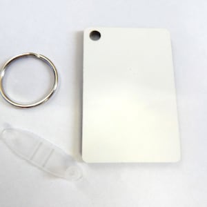 May include: A blank, rectangular white keychain tag with a hole at the top, a silver key ring, and a clear plastic attachment. The items are arranged on a white surface.