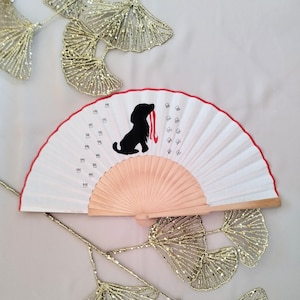 Puede incluir: Un abanico plegable blanco con una silueta negra de un perro con una cinta roja. El abanico tiene un mango de madera y está decorado con números negros.