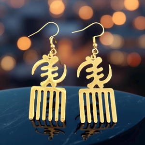 May include: A pair of gold earrings featuring the Adinkra symbol Sankofa, which represents the importance of learning from the past. The symbol is depicted as a bird with its head turned backward, holding an egg in its beak. The earrings are made of metal and have a comb-like design.