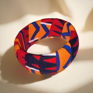 May include: A fabric-covered bangle bracelet with a vibrant geometric pattern. The design features red, orange, blue, and black shapes. The bracelet is a bold accessory, perfect for adding a pop of color to any outfit.
