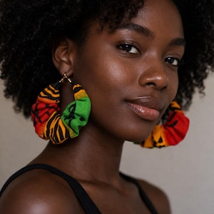 May include: A close-up portrait features a person wearing large, colorful hoop earrings. The earrings are made of fabric in red, yellow, green, and black patterns. The person is wearing a black tank top.