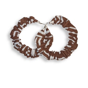 May include: A pair of hoop earrings with a brown and white zebra print fabric covering the hoops. The earrings are connected by a silver-colored clasp. The fabric is gathered, creating a textured appearance.