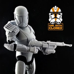 May include: A white plastic action figure of a Star Wars clone trooper holding a blaster rifle. The figure is standing on a black base. The figure is wearing a helmet with a visor. The figure is part of the One Sixth Clones line of action figures.