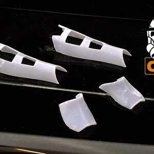 May include: Four white plastic pieces for a model kit. The pieces are shaped like wings and have a series of rectangular holes.