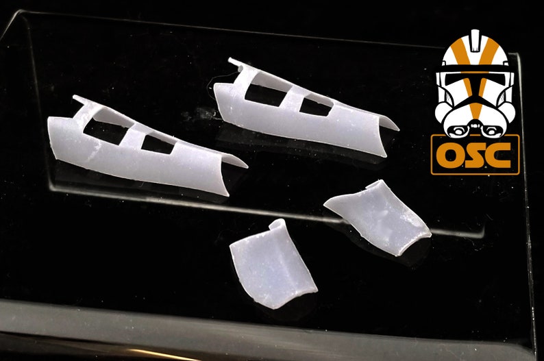 May include: Four white plastic pieces for a model kit. The pieces are shaped like wings and have a series of rectangular holes.