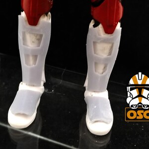 May include: A pair of white plastic action figure boots with a red and white design. The boots have a textured surface and are designed to look like armor.
