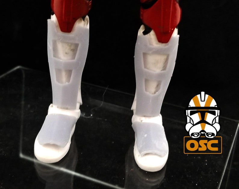 May include: A pair of white plastic action figure boots with a red and white design. The boots have a textured surface and are designed to look like armor.