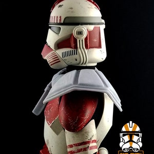 Puede incluir: Un casco de soldado clon de Star Wars blanco y rojo con un acabado de pintura desgastado y descolorido. El casco tiene una visera roja y una protecci&oacute;n de cuello gris. El casco es parte de una figura de acci&oacute;n a escala 1/6.
