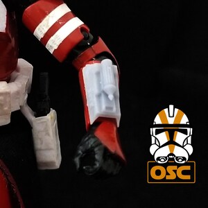 May include: A red and white action figure of a Star Wars clone trooper with a white blaster rifle on its hip and a white and gray accessory on its left arm.