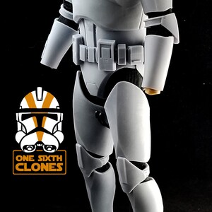 May include: A white plastic action figure of a Star Wars clone trooper. The figure is wearing a white helmet and armor with black accents. The figure is standing on a black base.