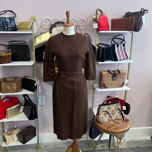 May include: A brown vintage dress with a matching belt is displayed on a mannequin. The dress has three-quarter sleeves and a knee-length skirt. Various vintage handbags are displayed on shelves in the background.