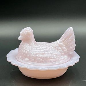 Op de afbeelding: Een lichtroze kipvormige schaal met een geschulpte rand. De kip zit op een nest en de schaal is van glas. De kip heeft gedetailleerde veren en een getextureerd oppervlak. De schaal wordt waarschijnlijk gebruikt om te serveren of tentoon te stellen.