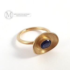 May include: A gold ring with a blue sapphire set in a textured, oval-shaped setting. The ring is made by MÜLLER COLLECTION Schmuckmanufaktur.