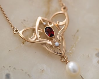 May include: A gold necklace with a pendant featuring a red gemstone surrounded by smaller white gemstones. The pendant is in the shape of a stylized heart with a single pearl dangling from the bottom.