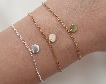May include: Three delicate chain bracelets with small round charms. The bracelets are layered on a person's wrist. The top bracelet has a gold charm, the middle bracelet has a silver charm, and the bottom bracelet has a silver charm.