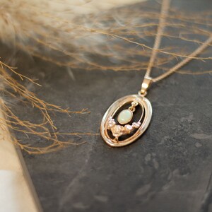May include: A gold necklace with a delicate oval pendant featuring a white opal gemstone surrounded by smaller stones. The pendant is set in a gold frame with a detailed design.