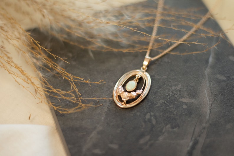May include: A gold necklace with a delicate oval pendant featuring a white opal gemstone surrounded by smaller stones. The pendant is set in a gold frame with a detailed design.
