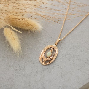 May include: A gold necklace with a delicate oval pendant featuring a white opal stone surrounded by smaller stones. The necklace is on a gray background.