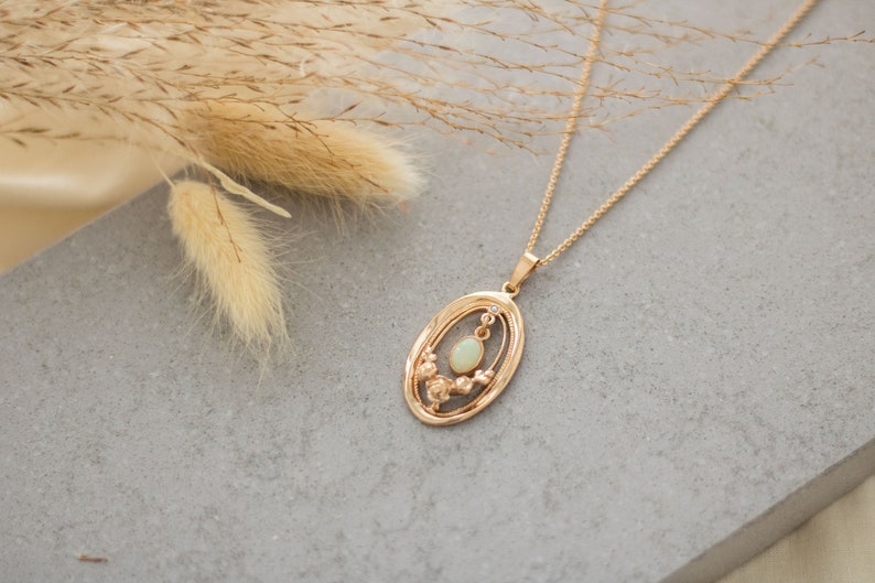 May include: A gold necklace with a delicate oval pendant featuring a white opal stone surrounded by smaller stones. The necklace is on a gray background.