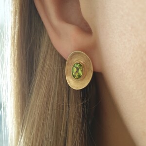 May include: A pair of gold earrings with a green gemstone in the center of each earring. The earrings are round and have a textured surface.