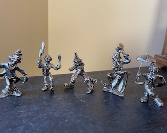 May include: A collection of five small, silver-colored clown figurines in various poses. One clown juggles clubs, another balances a hat, and another holds an umbrella. The figurines are detailed and appear to be made of metal.