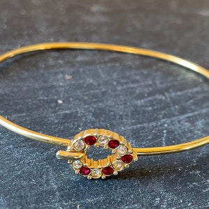 May include: A gold bangle bracelet with a red and clear rhinestone wreath charm.